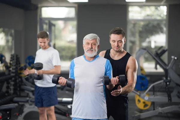 Trainer helping to work out elderly man with dumbbells. — Stock Photo, Image