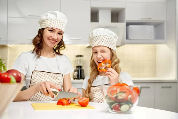 Mom cutting vegetables, daughter biting pepper slice.