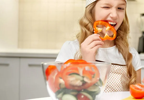 Lustiges Mädchen beißt Paprikascheibe, kocht Salat. — Stockfoto