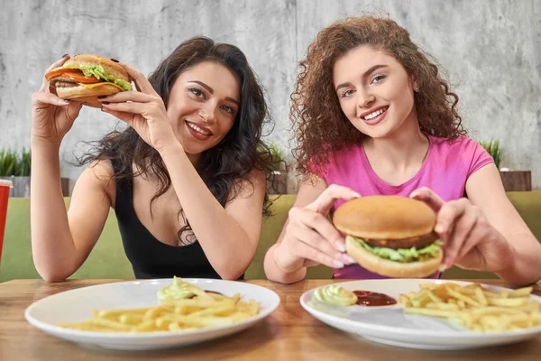 Zwei schöne Mädchen sitzen im Café und halten Hamburger in der Hand. — Stockfoto