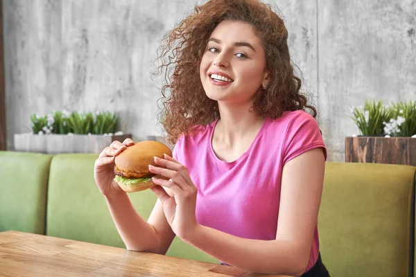 Mädchen sitzt im Café, hält leckeren Hamburger in der Hand und lächelt. — Stockfoto