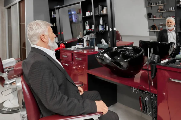 Ancien homme portant un costume intelligent noir assis dans le salon de coiffure — Photo