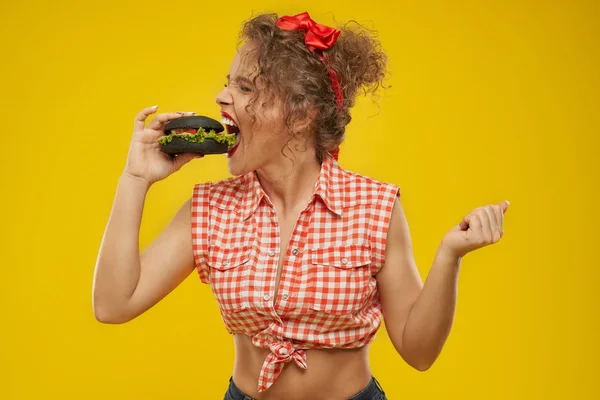Modell mit lockigem Haar rote Schleife beißt Hamburger. — Stockfoto