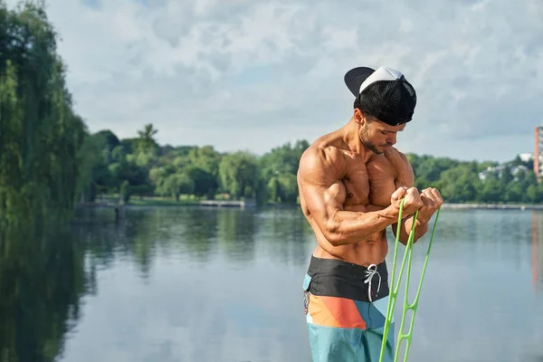 Masculino usando expansor y haciendo ejercicios deportivos cerca del agua . — Foto de Stock