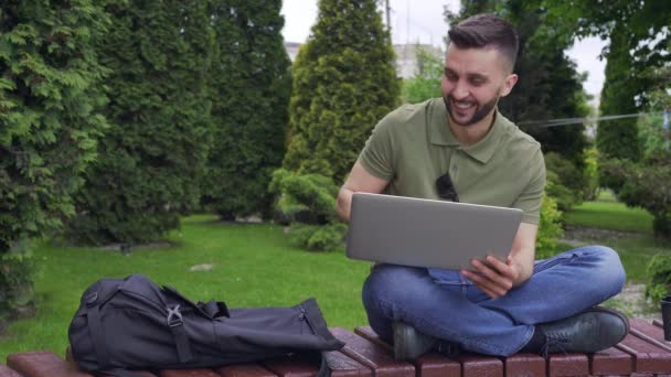 Счастливый бородатый мужчина отдыхает с ноутбуком в зеленом городском парке — стоковое видео