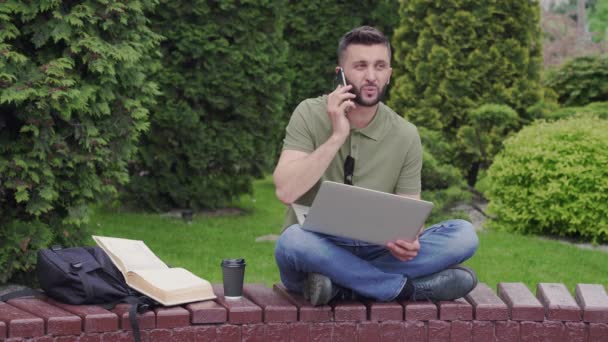 Schöner Mann mit Laptop und Handy sitzt im Park — Stockvideo