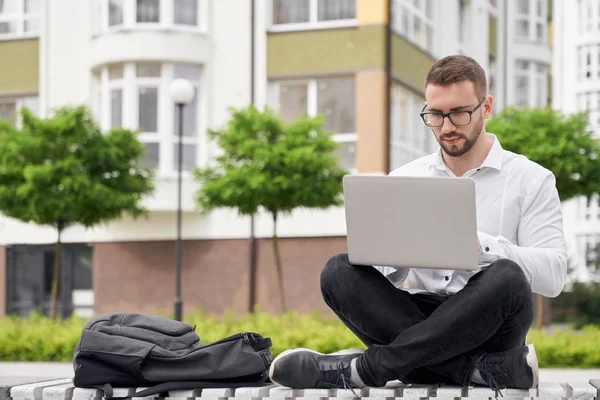 Homem de óculos no banco segurando laptop de joelhos, trabalhando . — Fotografia de Stock