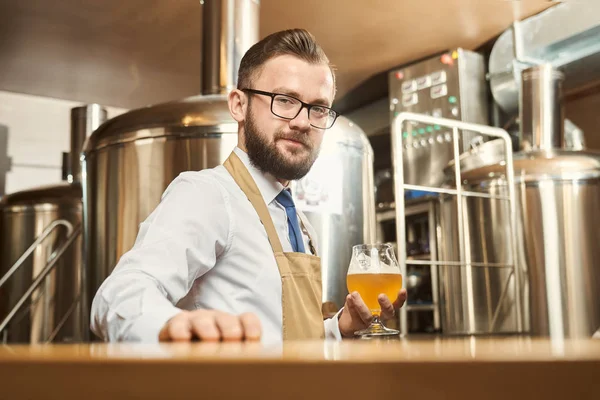 Cervecero joven sonriendo mientras mantiene el vaso de cerveza de oro — Foto de Stock