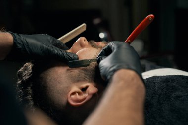 Handsome bearded man is getting shaved by hairdresser clipart