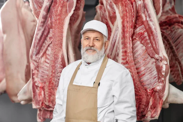 Carniceiro profissional em uniforme de pé perto de carcaças de porco — Fotografia de Stock