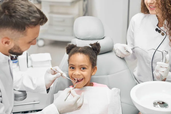 Chica sentada en la silla dental sonriendo mientras los dentistas trabajan —  Fotos de Stock