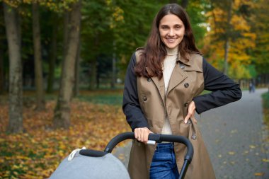 Gülümseyen bir kadın bebek arabasıyla parkta duruyor..