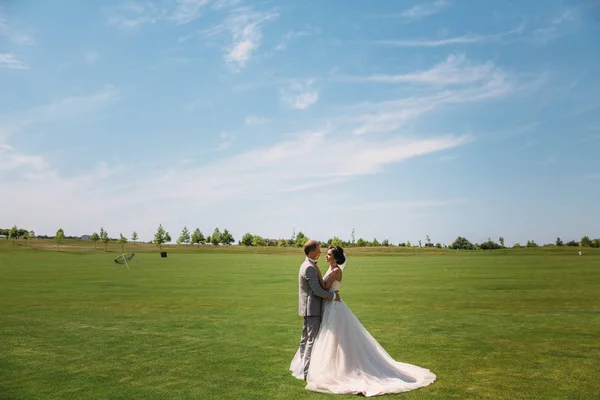 Молодята гуляють уздовж зеленого поля гольф-клубу в день весілля. Наречений у діловому костюмі сірий, а наречена в розкішній білій сукні з вуаллю тримається за руки . — стокове фото