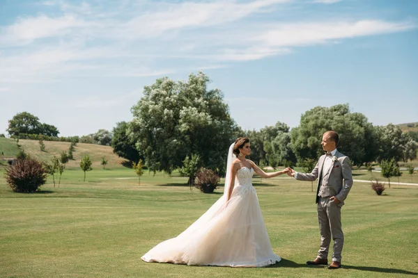 Frischvermählte spazieren an einem Hochzeitstag über die grüne Wiese des Golfclubs. Der Bräutigam im Businessanzug ist grau und die Braut im weißen Luxuskleid mit Schleier hält Händchen. — Stockfoto