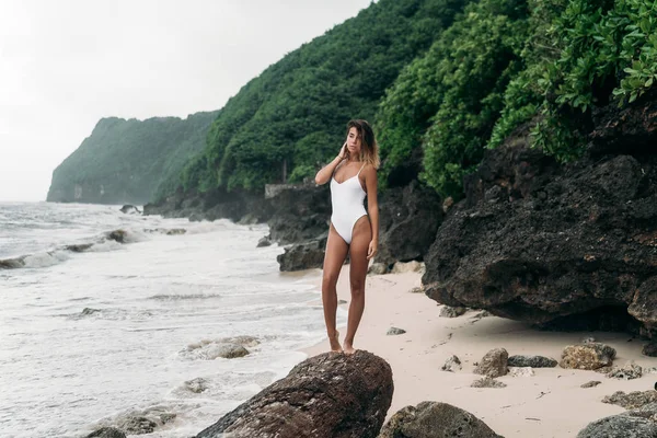 Sexy dziewczyna z sexy ciało i kręcone włosy. Uroczą kobietą w biały strój kąpielowy w pobliżu skały na plaży. Modelu pozowanie na letnie wakacje na wyspie, z niesamowitym widokiem. — Zdjęcie stockowe