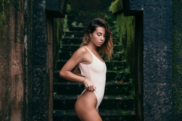 Amazing girl in a beige swimsuit on the background of a black stone wall with a chariot. Beautiful model with sexy body and curly hair posing outdoors. — Stock Photo, Image