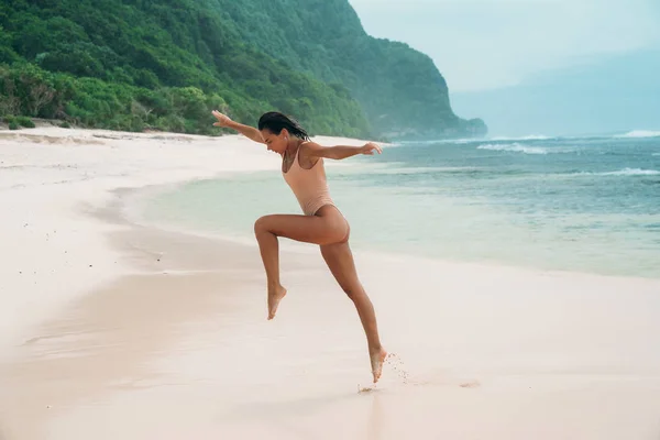 Uma mulher corre ao longo da praia com areia branca. Atleta feminina está envolvida em esportes pela manhã perto do oceano na ilha. Modelo em um maiô está pulando . — Fotografia de Stock