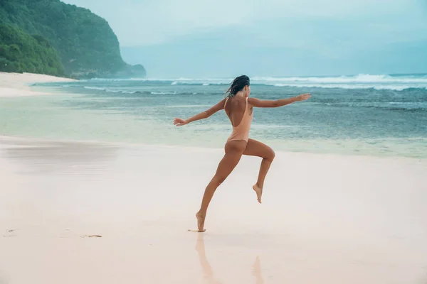 Uma mulher corre ao longo da praia com areia branca. Atleta feminina está envolvida em esportes pela manhã perto do oceano na ilha. Modelo em um maiô está pulando . — Fotografia de Stock