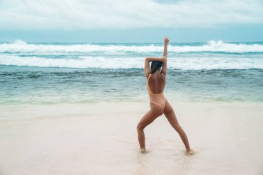 Kız sahilde su yoga yapıyor. Güzel seksi bir vücut spor yaparken bir mayo modeliyle açık havada okyanus yakın uygular. Genç kadın tatile güneşlenme.