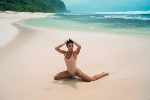 The model in a sexual pose kneels on a white beach against a background of blue waves and green rocks. A girl in a bathing suit posing on a white empty beach. — Stock Photo, Image