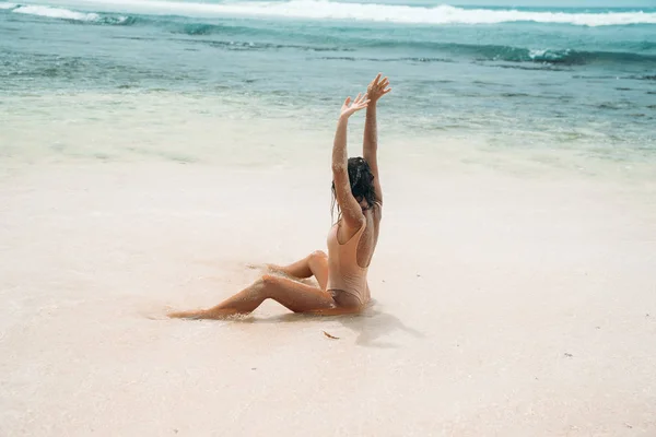 Ein schönes Mädchen im Bikini sitzt am Strand und zieht ihre Hände in die Sonne. das Modell bräunt am Ufer des blauen Ozeans. — Stockfoto