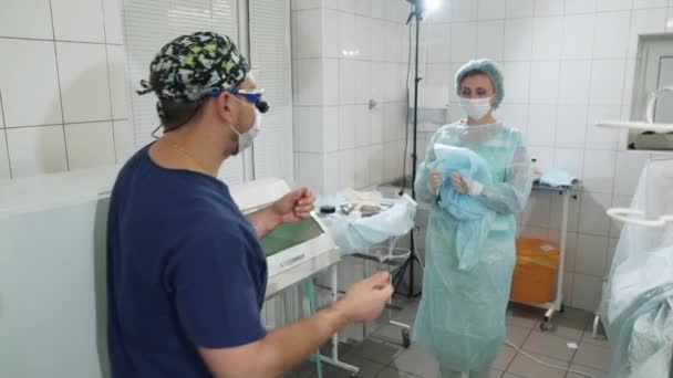 El doctor entra al quirófano. Una enfermera ayuda a vestir una bata de médico en el quirófano. Preparándose para la cirugía. Sala de operaciones en el hospital. Una enfermera y un cirujano están empezando a — Vídeos de Stock