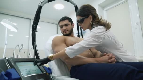 Une femme esthéticienne comprend un dispositif pour la procédure d'épilation du corps d'un homme. Elle appuie son doigt sur l'écran tactile. Le patient s'allonge sur la chaise, le médecin est prêt pour — Video