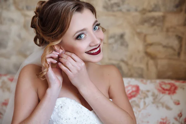 Uma menina agradável em um vestido de noiva e véu coloca brincos e sorrisos amplamente. A noiva com grandes olhos azuis, brancos até dentes e lábios vermelhos brilhantes . — Fotografia de Stock