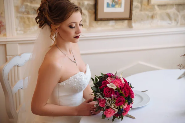 A noiva com cachos e lábios vermelhos senta-se em um salão brilhante e admira seu buquê brilhante de rosas vermelhas e rosa . — Fotografia de Stock