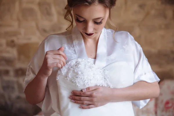 Uma menina com cachos e pestanas longas olha para baixo em seu vestido de noiva, que ela segura com a mão. A noiva admira o bordado, padrões e tecido de seu traje de casamento . — Fotografia de Stock