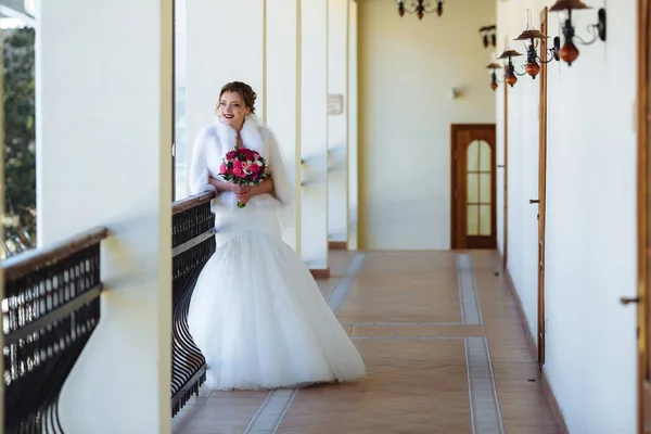 A noiva em um vestido branco com uma saia apertada está de pé na varanda e admirando a natureza. A menina está segurando um buquê de flores rosa . — Fotografia de Stock