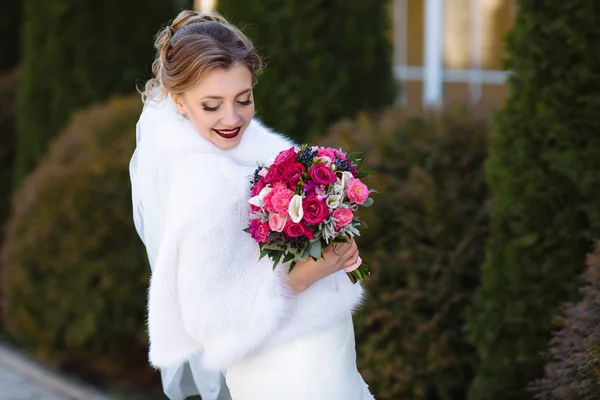 Nevěsta v bílém plášti pózuje venku, v ruce držel světlé kytice květin. Dívka se sklonil oči a ukázal její make-up přírodní tóny. — Stock fotografie