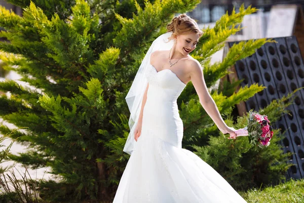Die Braut geht im Park zwischen den Tannen spazieren, hat Spaß und spielt mit ihrem Hochzeitsstrauß. schlanke lächelnde Mädchen in einem weißen Kleid. — Stockfoto