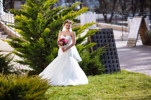 En flicka i en lång slöja och en mild vit klänning poserar mot grönskan i parken. Han håller i sin hand en ljus blommig bukett. — Stockfoto