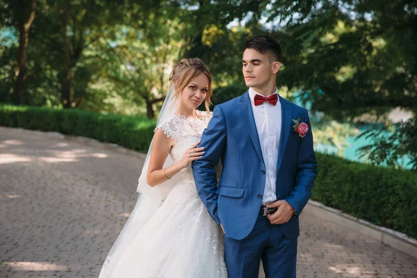 Um retrato de casamento de um belo casal. A noiva com o cabelo loiro reunido em um vestido de renda segura-se ao ombro corajoso do noivo em um terno azul em uma borboleta vermelha. O casal está em pé — Fotografia de Stock