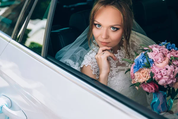 Uma loira de olhos grisalhos num vestido de noiva está sentada no carro. Retrato de uma menina bonita com maquiagem e um buquê de flores. O modelo está a olhar para a câmara . — Fotografia de Stock
