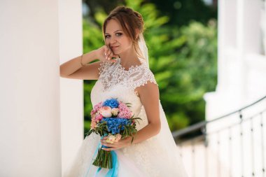 Bir gelinlik ince bir gelin gülümseyen ve parkta yeşil bir arka plan poz. Bir kız bir buket çiçek ile evli ve mutlu oluyor