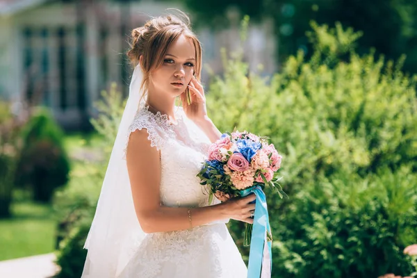 Hermosa novia delgada posando contra la vegetación en el parque. Ella está vestida con un hermoso vestido de novia blanco, en su mano sostiene un elegante ramo de flores y mira atentamente a la cámara . —  Fotos de Stock