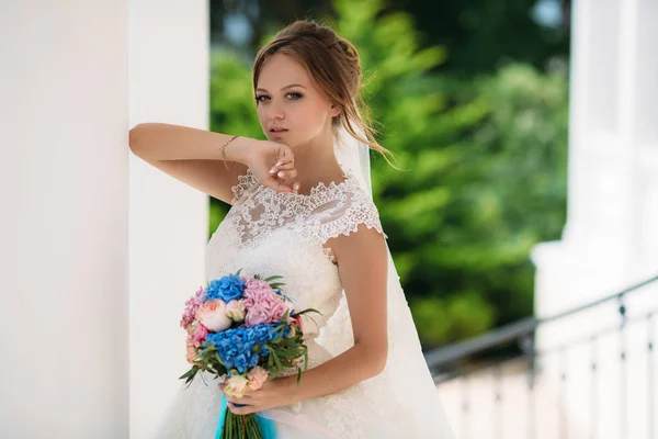 Noiva loira com olhos azuis posando em vestido de noiva com buquê de flores. A modelo levantou a mão para o rosto. O vento desenvolve as meninas cabelo loiro . — Fotografia de Stock