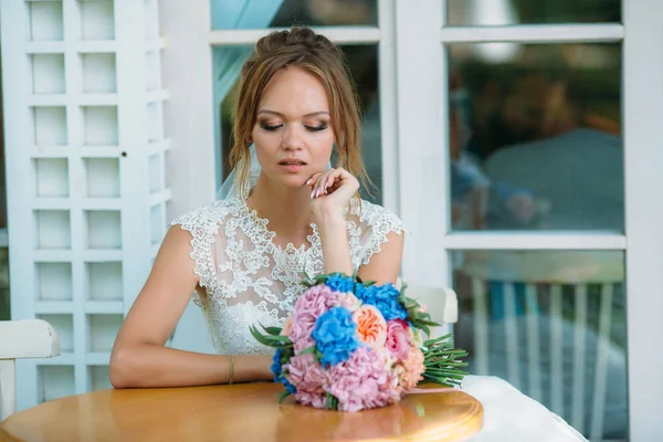 A menina no vestido de noiva senta-se à mesa sozinha e parece chateada. A noiva olha tristemente para um ramo de flores . — Fotografia de Stock