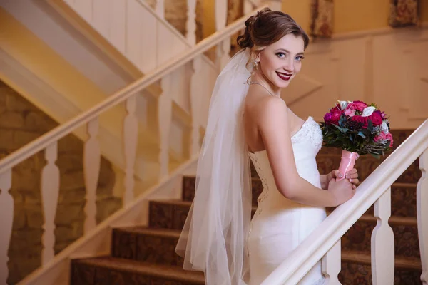 Blick von hinten schöne blonde Frau in Schleier und Brautkleid mit Blumenstrauß, lächelnd und die Treppe des großen Saals hinauf. — Stockfoto