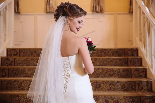 Blick von der Rückseite einer schönen Braut in einem Brautkleid mit Schnürung steigt die Treppe hinauf, dreht sich um und lächelt. Gesichtsmodell im Profil gedreht. — Stockfoto