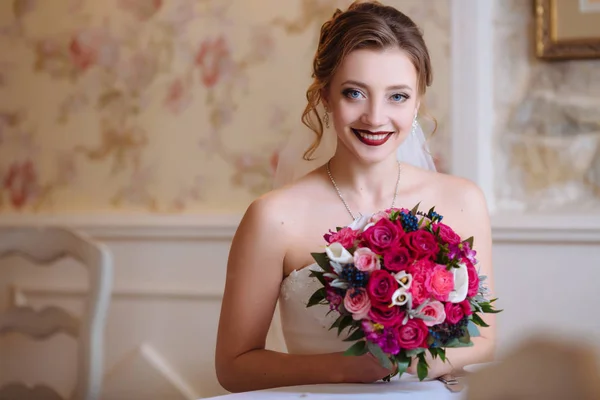 A noiva está segurando um buquê de flores vermelhas e sorrindo feliz sentado à mesa. Uma menina com olhos azuis e cachos se casa . — Fotografia de Stock