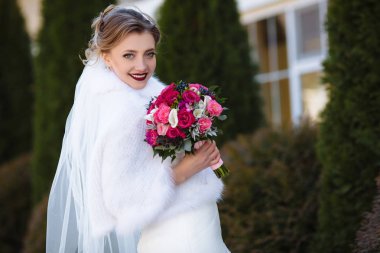 Gelin bir sıcak, sıcak pelerin açık havada uzun ağaçlar arka planı teşkil etmektedir. Kız gülümsüyor ve onu buket çiçek kokusu nefes.