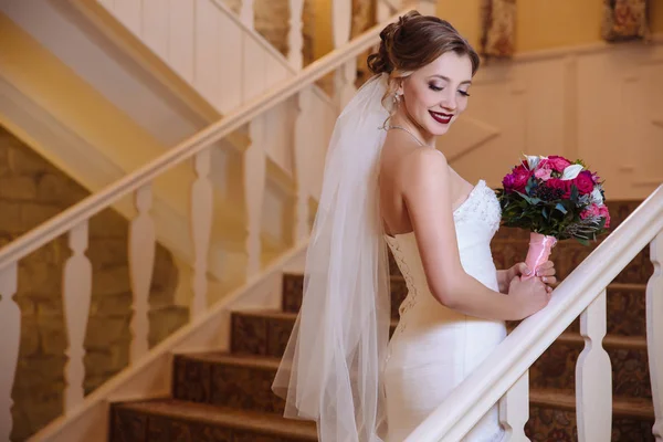 Uma bela noiva caminha ao redor do salão e sobe as escadas para seus convidados. A menina sorri e mostra seus belos dentes, admira um buquê de rosas vermelhas . — Fotografia de Stock