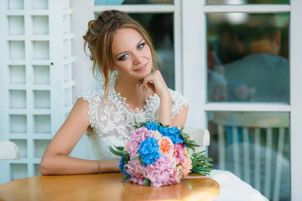 Die blonde Braut sitzt an einem Tisch und genießt einen schönen Tag. In der Nähe liegt ihr rosa-blauer Hochzeitsstrauß. die blauen Augen der Mädchen sagen, dass sie glücklich ist. — Stockfoto