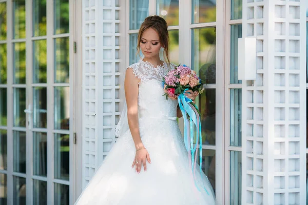 Die Braut posiert in einem Spitzenkleid in der Nähe der weißen Glasfenster. Make-up aus natürlichen Tönen. — Stockfoto