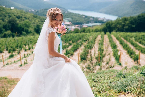 La novia se ríe y se regocija en el día soleado. La niña sostiene un elegante ramo de flores en sus manos y camina entre viñedos, campos y colinas. Admira el lago azul . —  Fotos de Stock