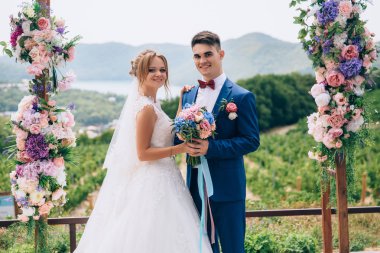 Güzel ve gülümseyerek gençlerin muhteşem bir doğanın ortasında bir çiçek kemerin altında poz olan. Bir kız bir gelinlik nişanlısının onun omuz üzerinden sarılmak. Bir kelebek ile bir takım elbise içinde bir iş adamı