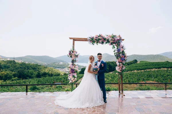 Nygifta är poserar i öppna luften nära blomma bågen och brett leende. Vackra par njuter utomhus av bröllopsdagen. — Stockfoto